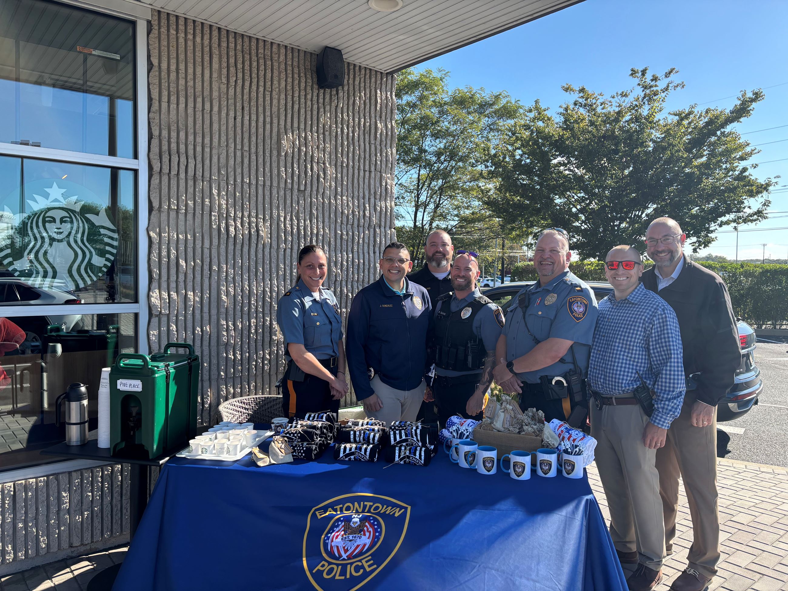 EatonTown Police staff pose outdoors by a merchandise table with mugs, cups, and folded items.